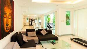 A modern living room with a sofa set, a glass coffee table, and a treadmill. - Villa Cheloni 3 Bedrooms (Kamala)