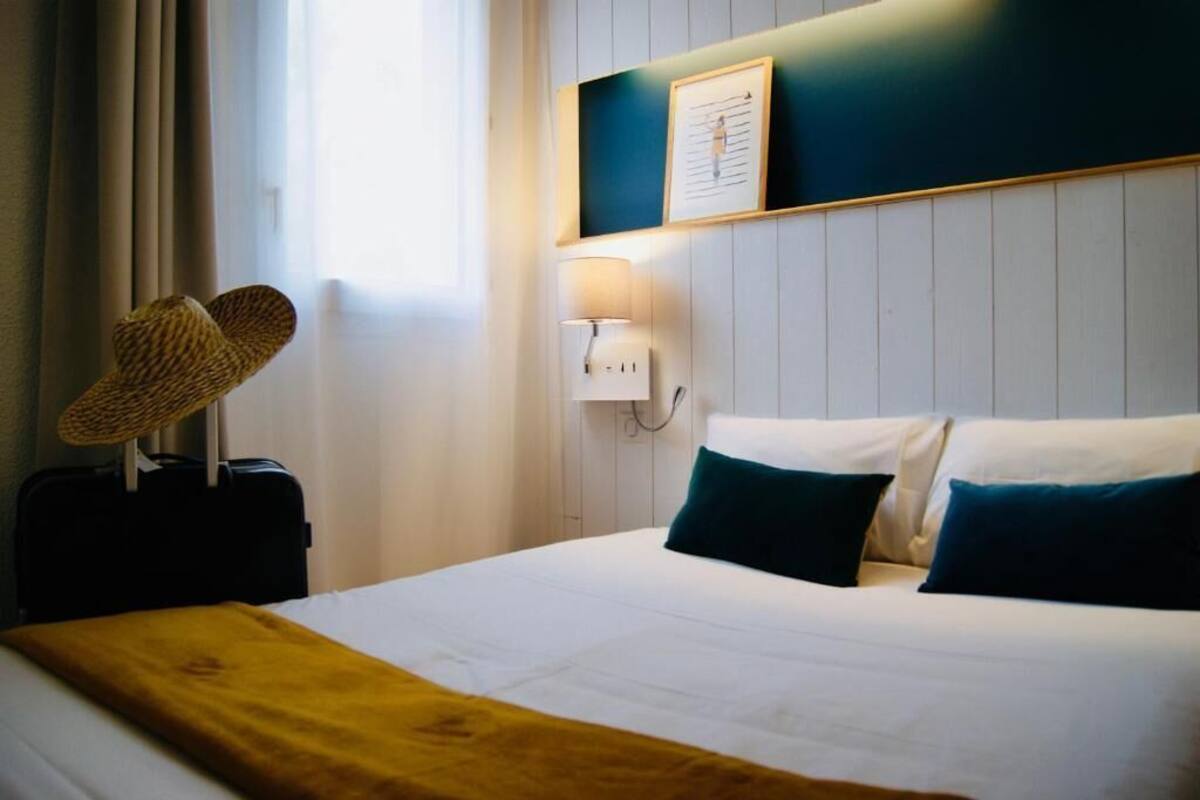 Une chambre d’hôtel avec un lit, une lampe, une valise et un chapeau accroché au mur.