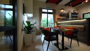 A modern dining area with a wooden table, black chairs, and orange cushions. There is a large window with curtains, a potted plant, and a bar area with shelves and bottles. - Manor Le Rayon Vert (Ji'an)