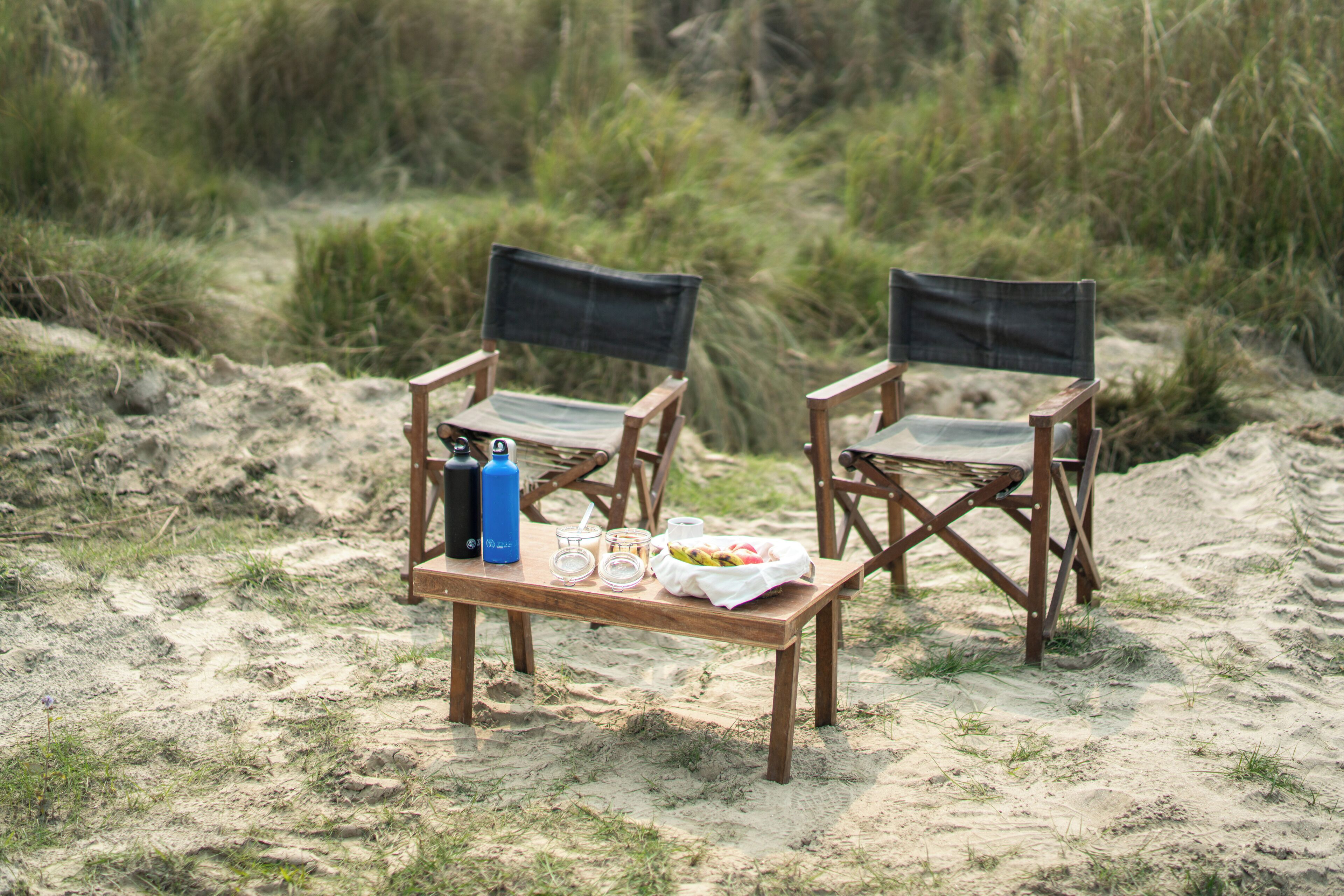 Dos sillas de madera con cojines negros sobre una playa arenosa con una mesa en medio.