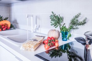 Una encimera de cocina con un lavabo, tabla de cortar, vaso de agua, tazón de tomates, una calabaza, una planta y una bolsa de pasta.