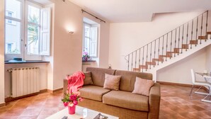 A living room with a sofa, a coffee table with a remote control and book, a staircase, and a window with a view of the outside. - Maison Palazzo Correale (Sorrento)