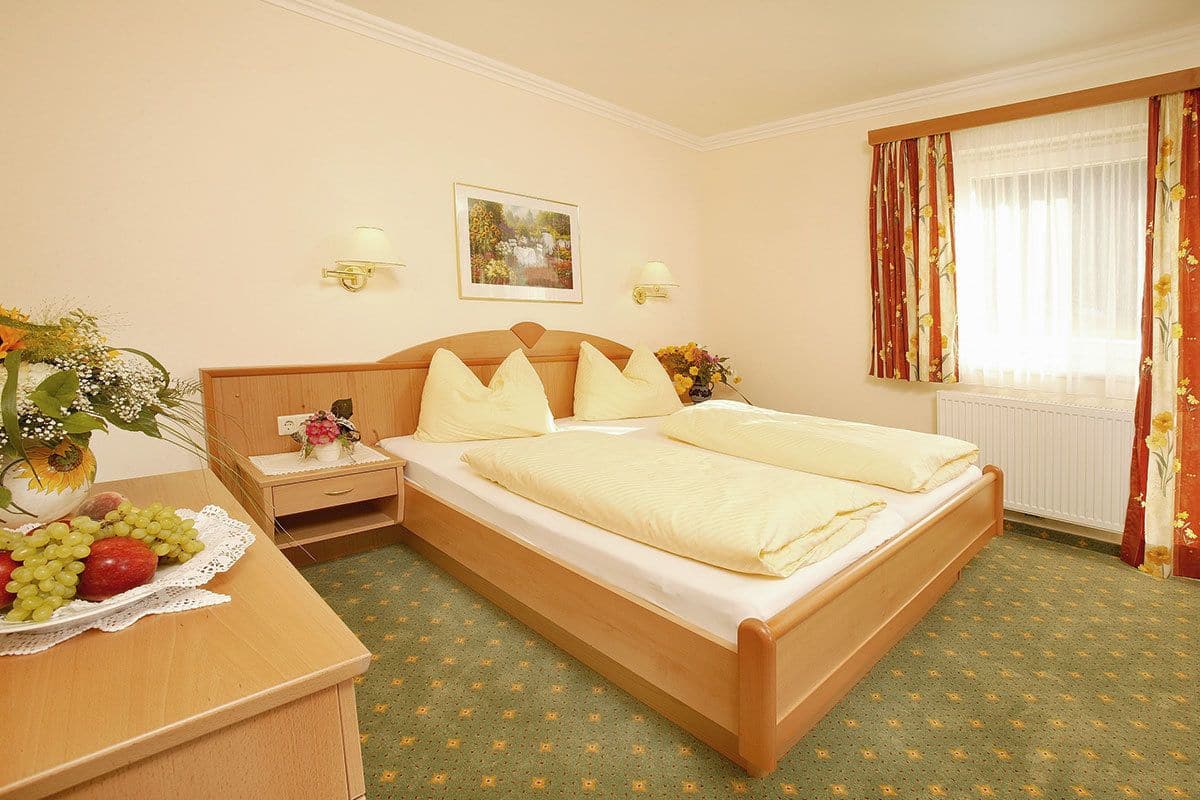 Une chambre d’hôtel avec un lit en bois, deux lampes de chevet, une table de chevet ornée d’un vase de fleurs et un panier de fruits.