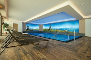 A modern indoor pool area with wooden flooring, lounge chairs, and a mural of a mountainous landscape. - Hotel Taxerhof (Radstadt)
