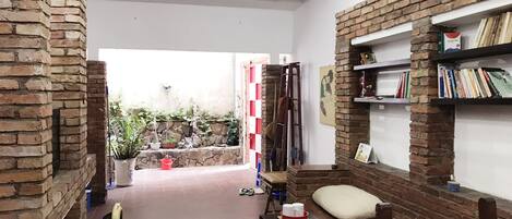 A living room with a brick fireplace, a bookshelf, a small table with a cushion, and a view into the garden.