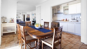 Un coin repas avec une table et des chaises en bois, un saladier rempli de fruits, et une cuisine en arrière-plan.