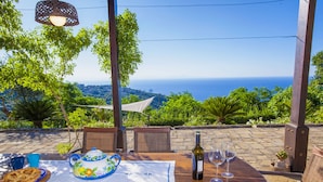 Une table dressée avec une théière, une bouteille de vin et des verres, offrant une vue panoramique sur la mer et une végétation luxuriante.