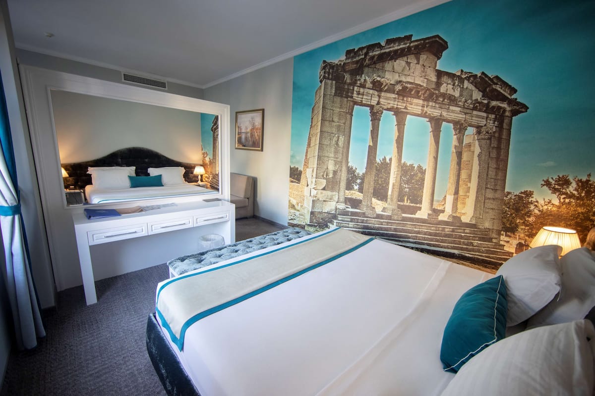A hotel room with a large bed, a desk, and a mural of ancient ruins.