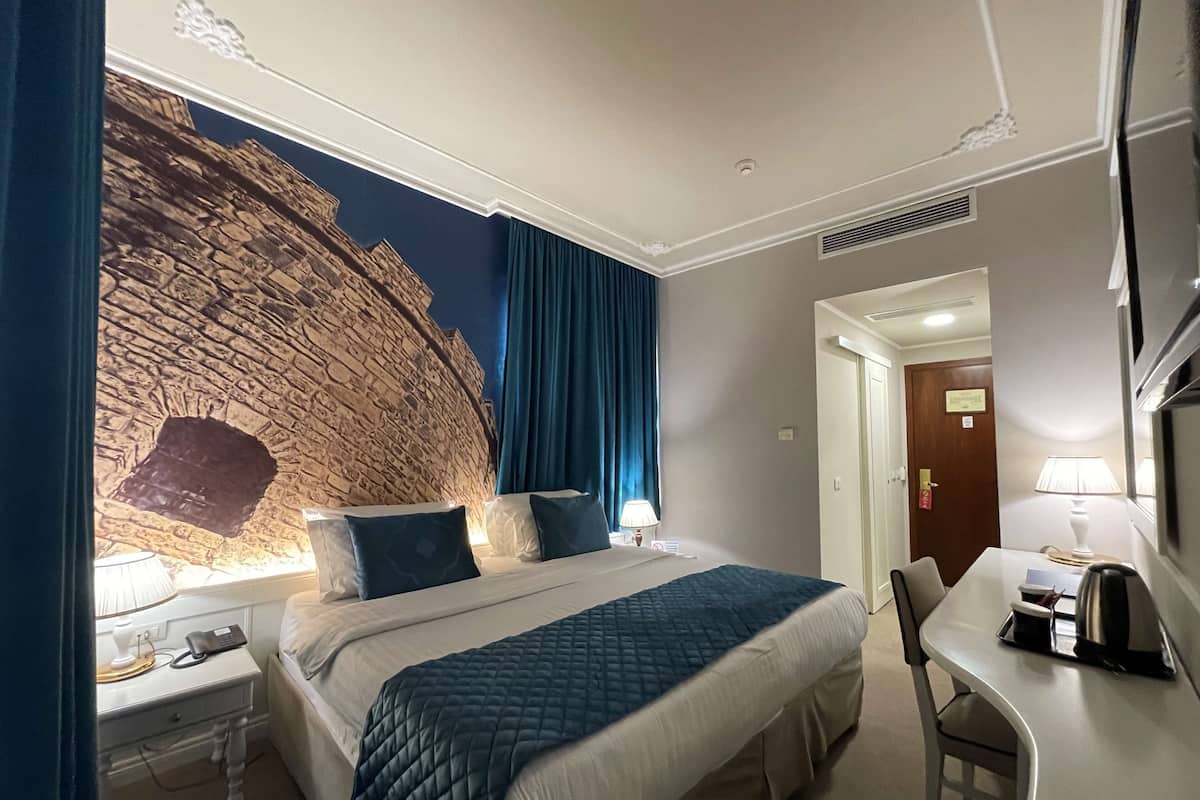 A hotel room with a large bed, a desk with a chair, a lamp, and a wall mural.