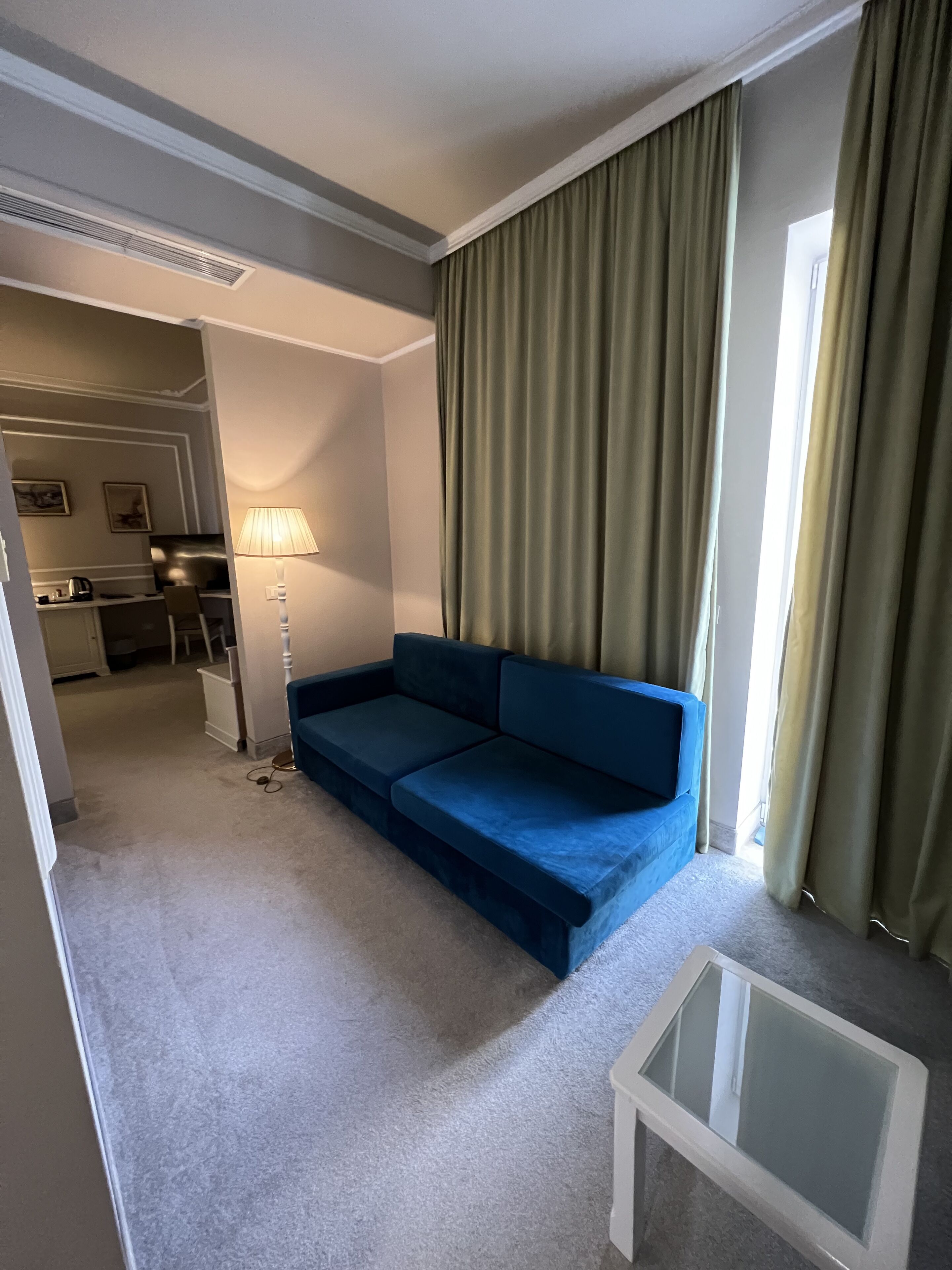 A compact living room with a blue sofa, a small glass table, and a lamp. A kitchen area is visible in the background.