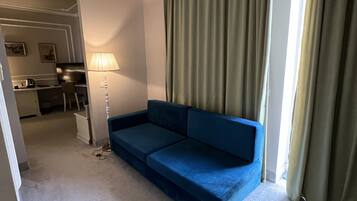 A compact living room with a blue sofa, a small glass table, and a lamp. A kitchen area is visible in the background.