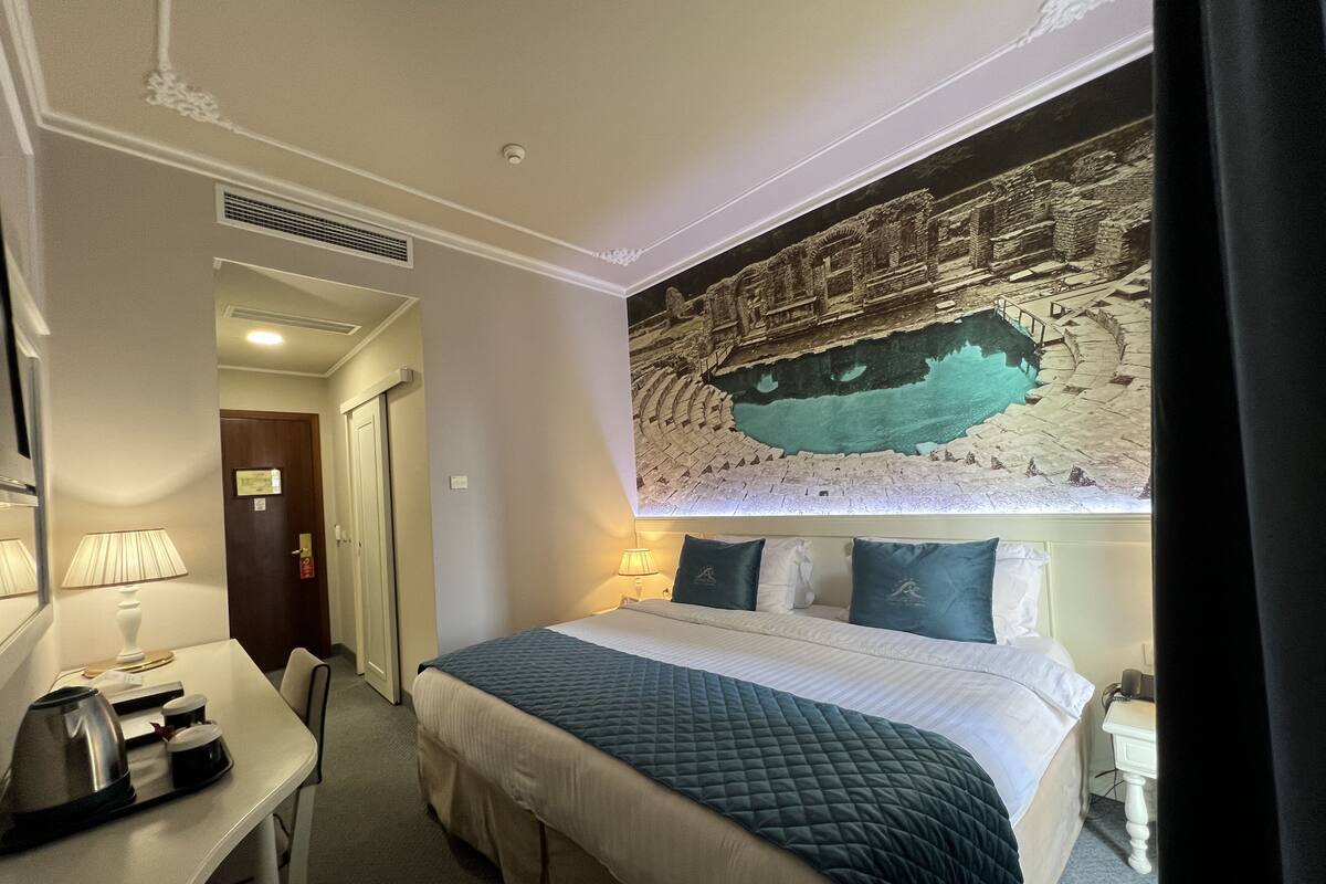 A hotel room with a bed, bedside table, lamp, and a mural of an ancient temple.