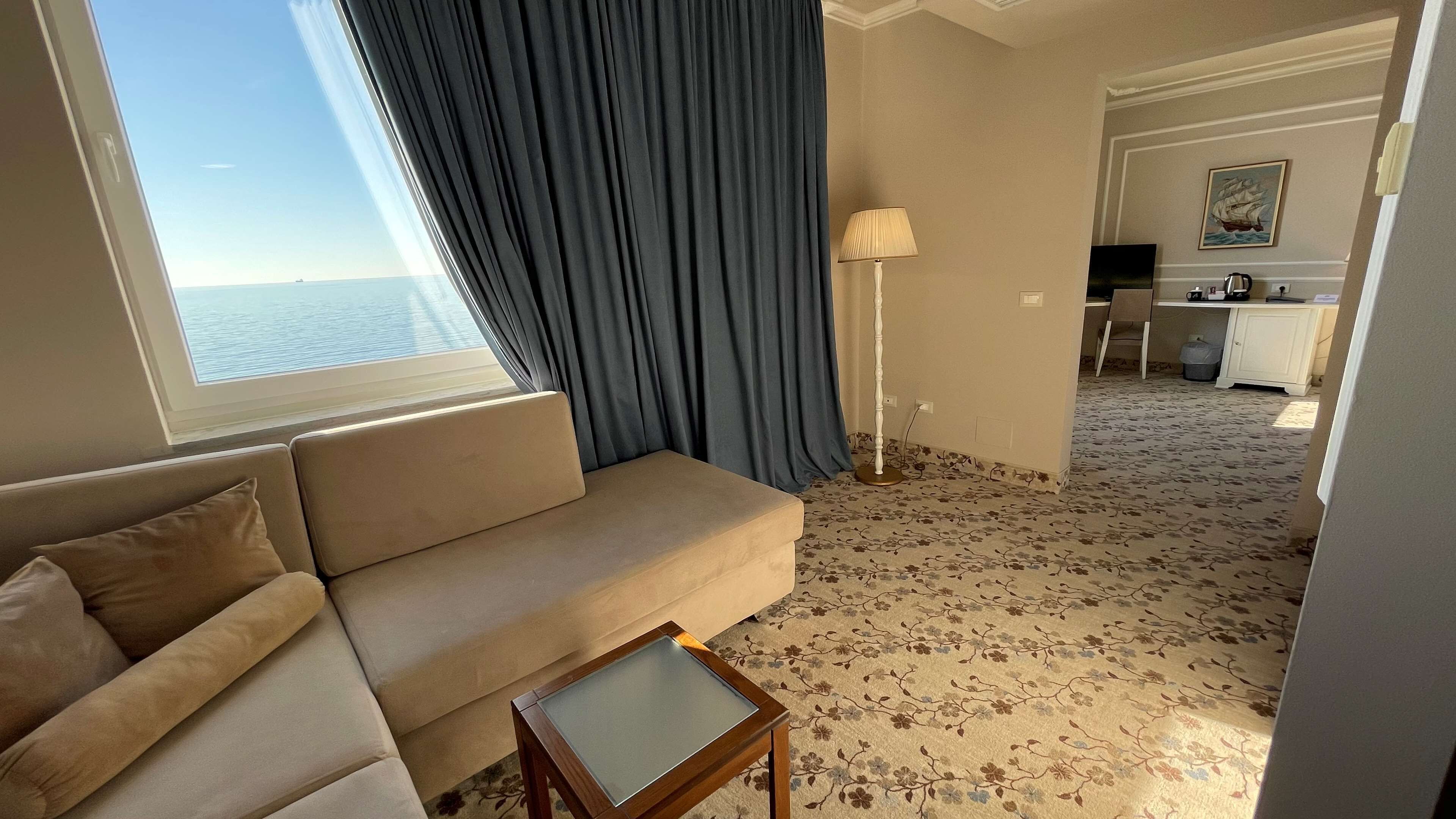 A hotel room with a sofa, a small table, a lamp, and a view of the sea through a window with curtains.
