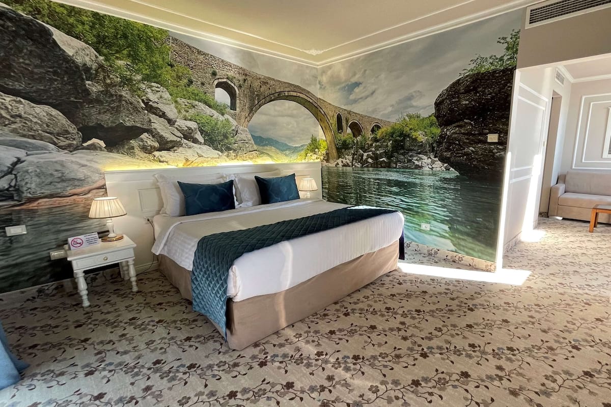 A bedroom with a large bed, a nightstand, a lamp, and a mural of a bridge over water.