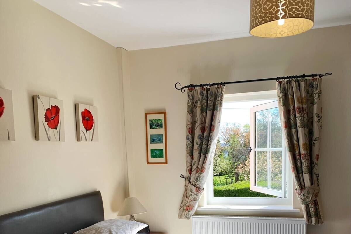 A bedroom with a bed, a nightstand, a lamp, a window with curtains, and floral artwork on the wall.
