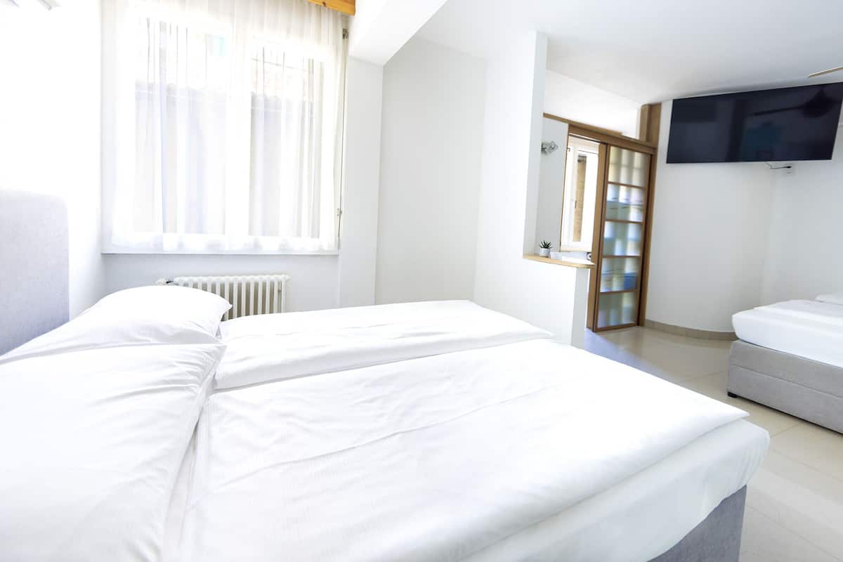 A hotel room with a bed, a TV mounted on the wall, a radiator, and a window with sheer curtains.