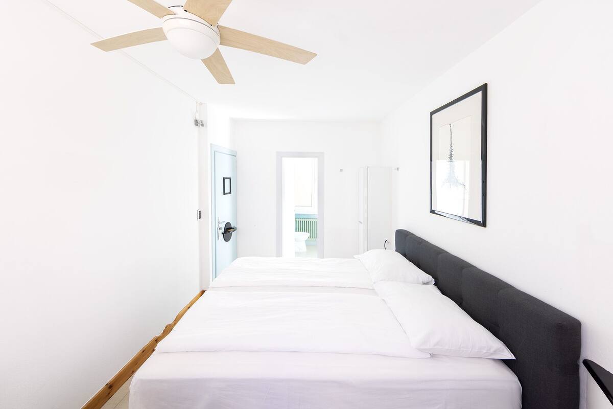 A hotel room with a bed, a ceiling fan, a framed picture on the wall, and a door leading to another room.