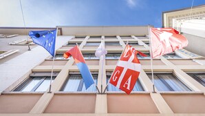 A building facade with multiple flags, including a blue flag with stars, a red and white flag, and a flag with the letters "DGA". - Hotel San Carlo (Lugano)