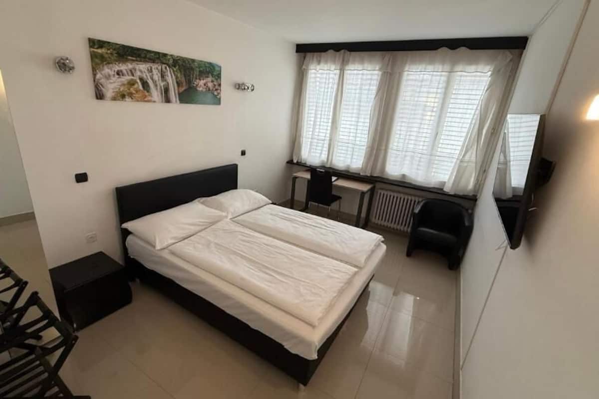 A bedroom with a bed, a desk, and a chair. There is a window with curtains and a painting on the wall.