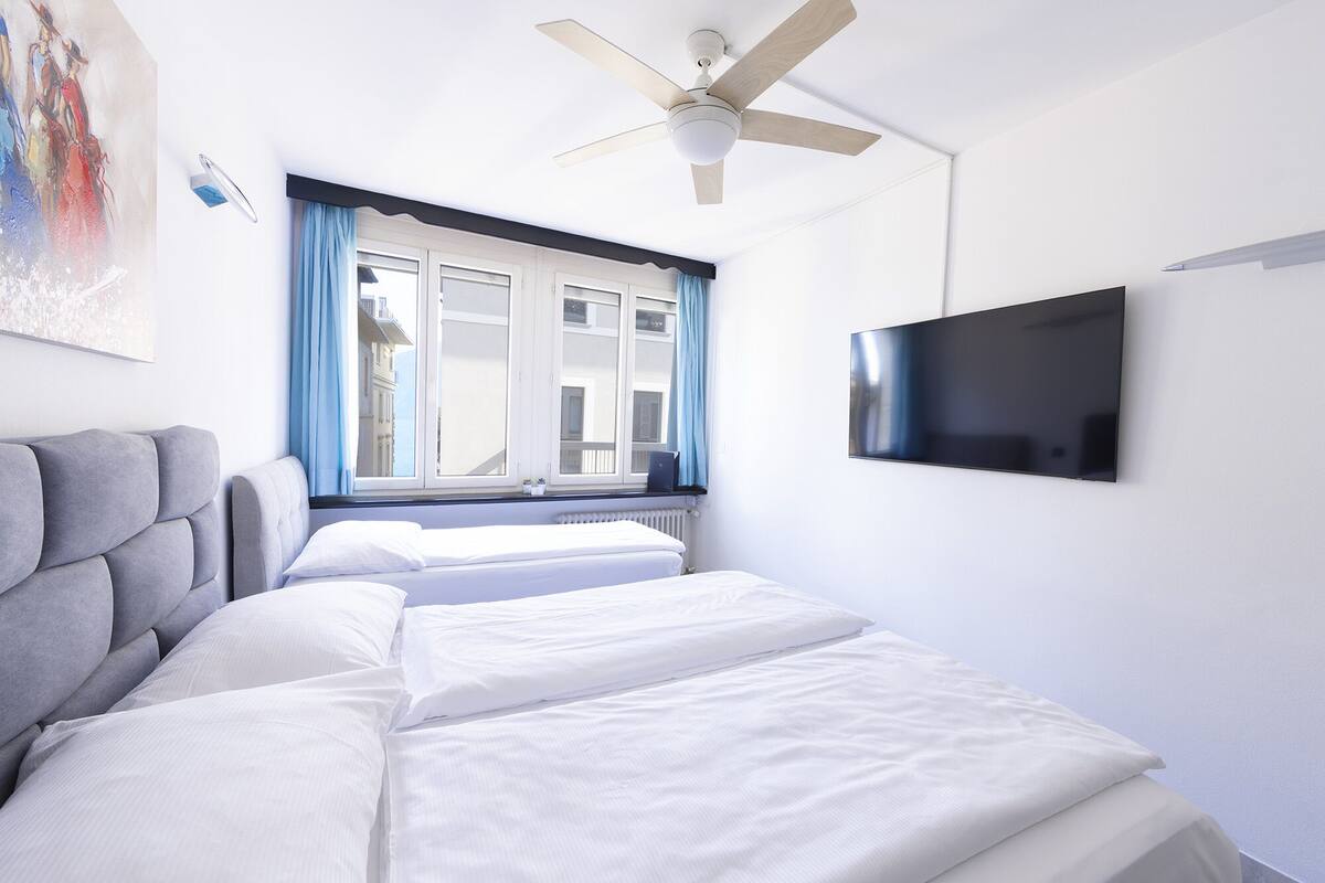 A hotel room with a bed, a ceiling fan, a window with curtains, and a flat-screen TV mounted on the wall.
