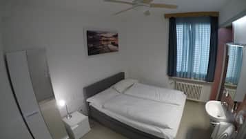 Ein Schlafzimmer mit Bett, Nachttisch, Deckenventilator, Fenster mit Vorhängen und Heizkörper, sowie ein Badezimmer mit Waschbecken und Spiegel.