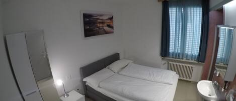 A bedroom with a bed, a nightstand, a ceiling fan, a window with curtains, a radiator, and a bathroom with a sink and mirror.
