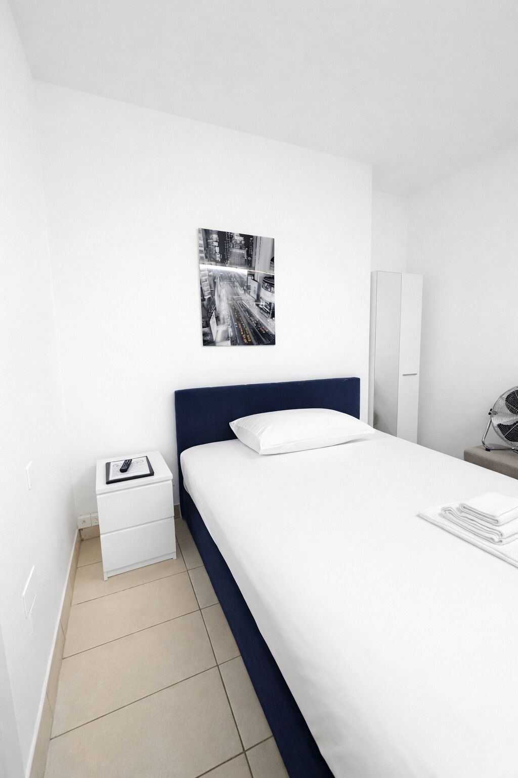 Moderni hotellihuone, jossa on siististi pedattu sänky, pieni yöpöytä, jossa on kaukosäädin, seinälle ripustettu taulu ja peili.