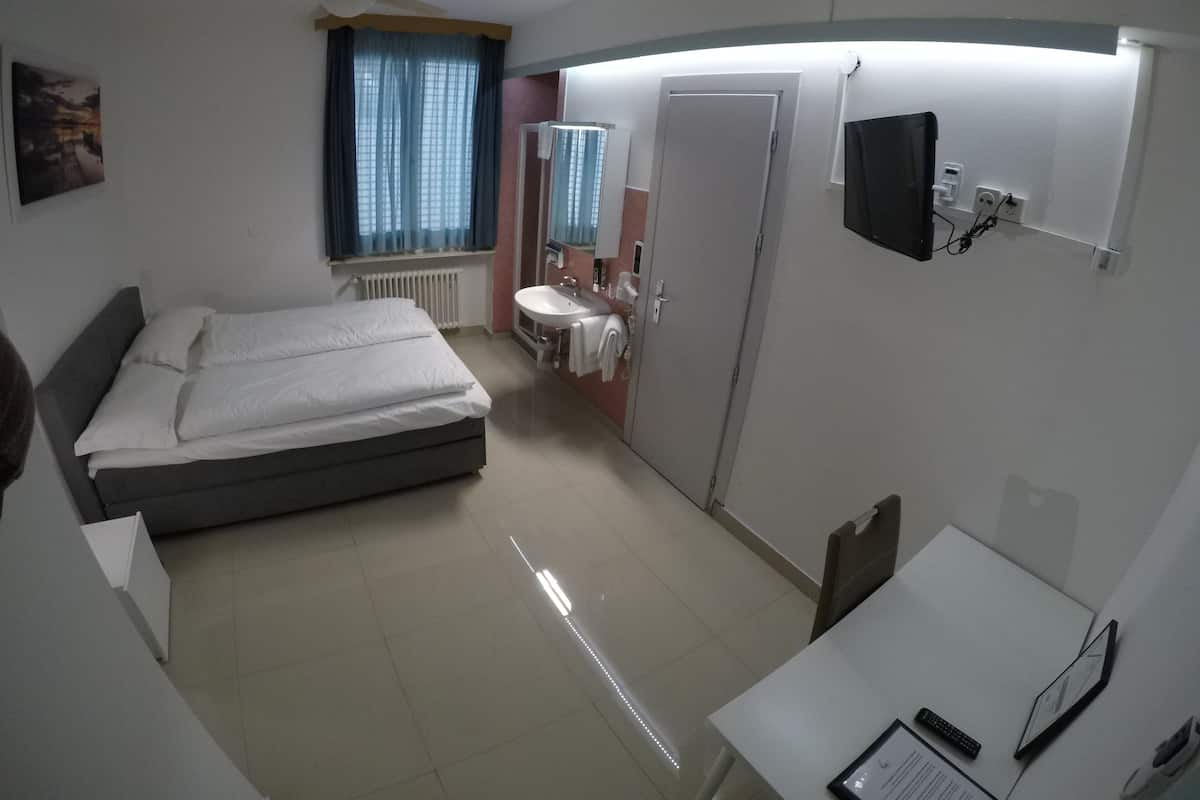 A hotel room with a bed, a desk, a chair, a window with curtains, a radiator, a sink, a mirror, and a ceiling fan.