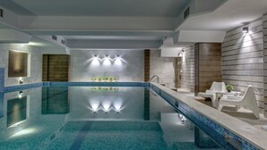 Una piscina interior moderna amb una superfície d'aigua cristal·lina, una zona d'estar integrada amb cadires blanques i una paret amb il·luminació integrada.
