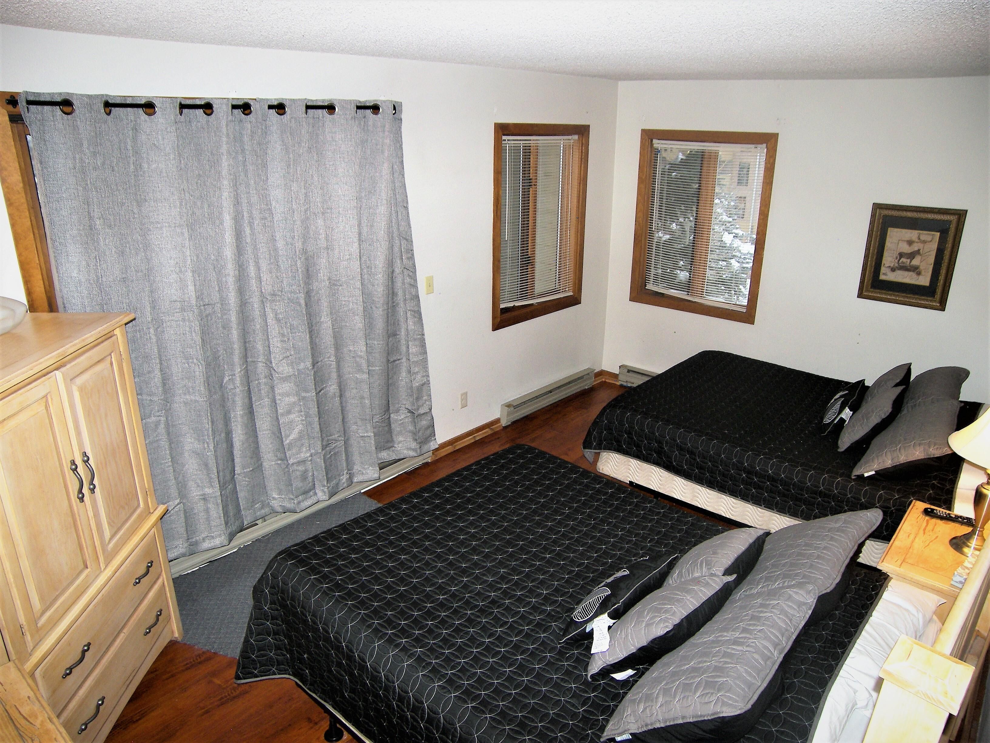 A bedroom with two beds, a wooden dresser, a window with blinds, and a curtain.