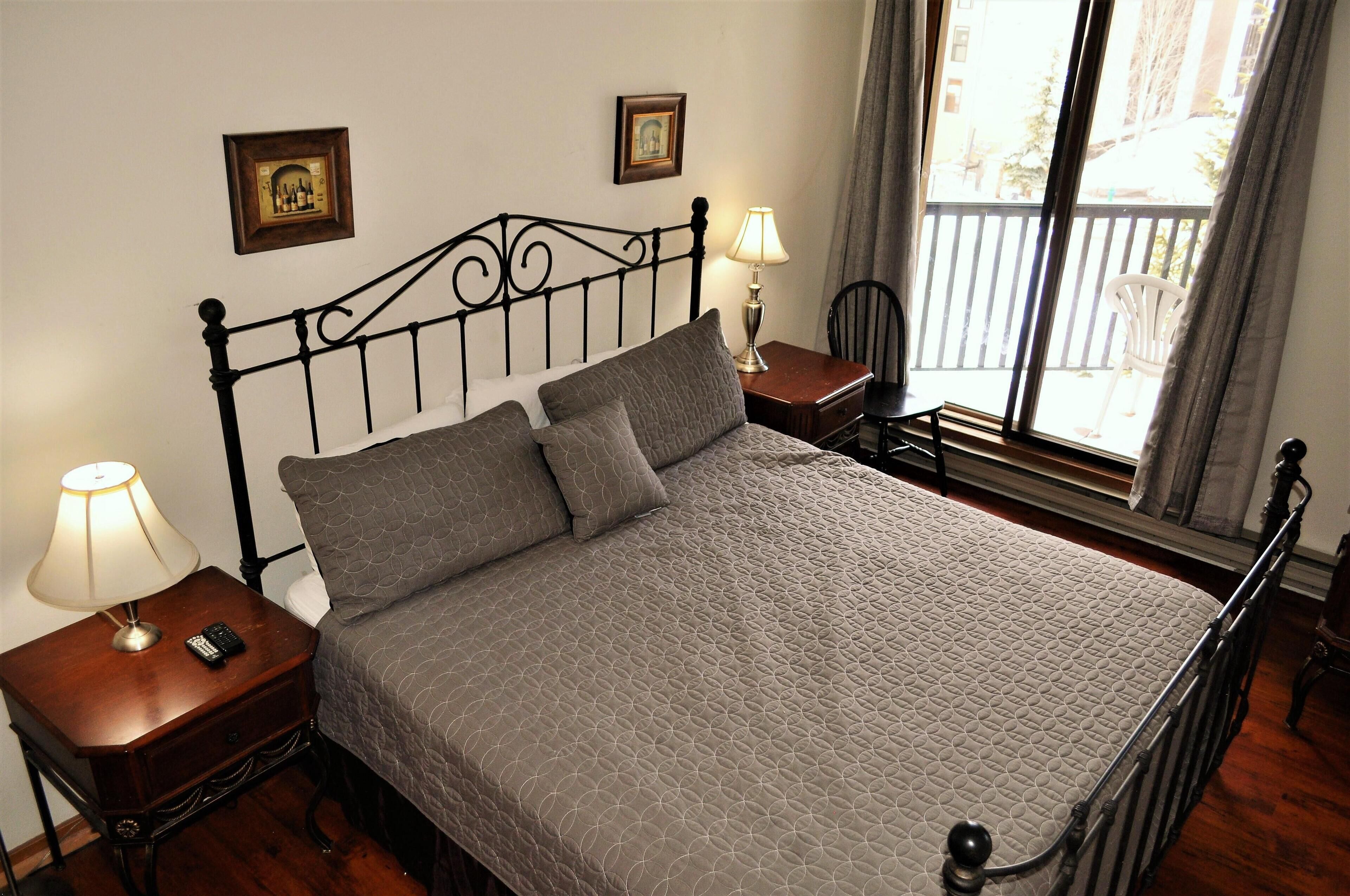 A neatly made bed with a quilted cover, a bedside table with a lamp, and a chair by the window.