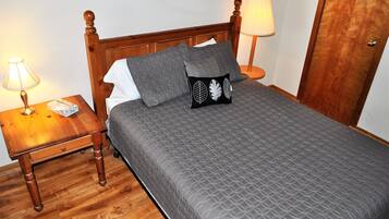 Ein ordentlich gemachtes Bett mit einer grauen Tagesdecke, zwei grauen Kissen und einem schwarzen Kissen mit Blättermotiv. Ein Holznachttisch mit Lampe steht daneben, im Hintergrund ist eine Holztür zu sehen.