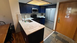 A compact kitchen with dark blue cabinets, a stainless steel refrigerator, a microwave, and a sink with a granite countertop. - Fox Pine Lodge 3 Bed 3 Bath (Copper Mountain)