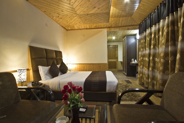 A hotel room with a large bed, a wooden ceiling, a desk with a lamp, a vase with flowers, and a curtain with a leaf pattern. - Victory Resorts (Manali)