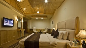 Habitación de hotel con techo de madera, una cama grande con almohadas, una mesita de noche con lámpara, un televisor en la pared y una pequeña mesa con sillas.