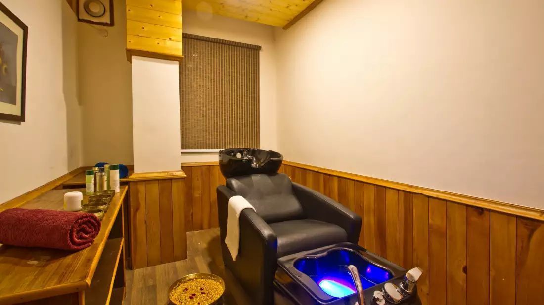 A massage chair with a heated base, a wooden bench with a red towel, and a small table with bottles.