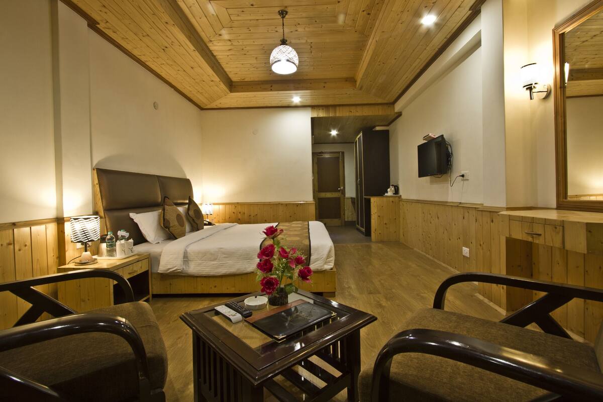 A hotel room with a wooden ceiling, a bed with a headboard, a bedside table with a lamp, a small table with a vase of flowers, and a mirror on the wall.