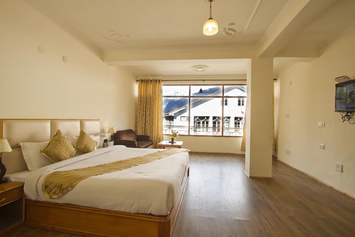 A hotel room with a large bed, a wooden floor, a window with a view of a building, and a television on the wall.