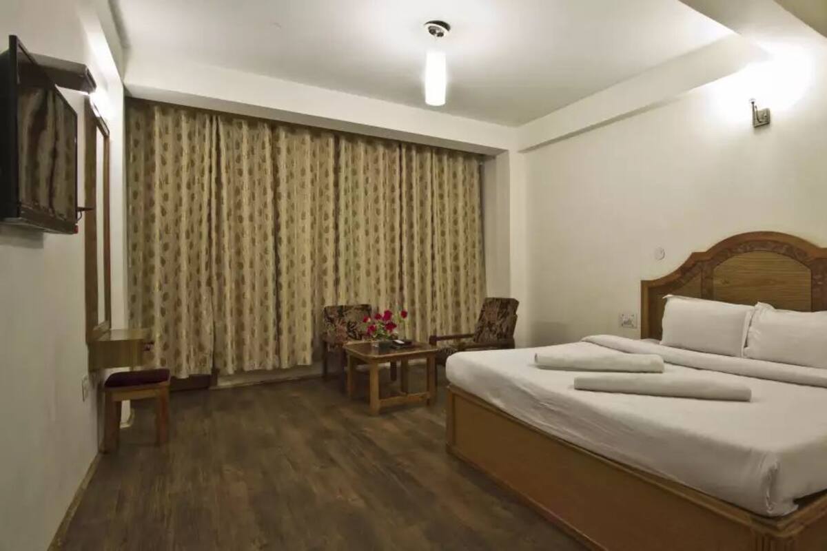 A hotel room with a large bed, a wooden headboard, a small table with a vase of flowers, a chair, and a curtain-covered window.