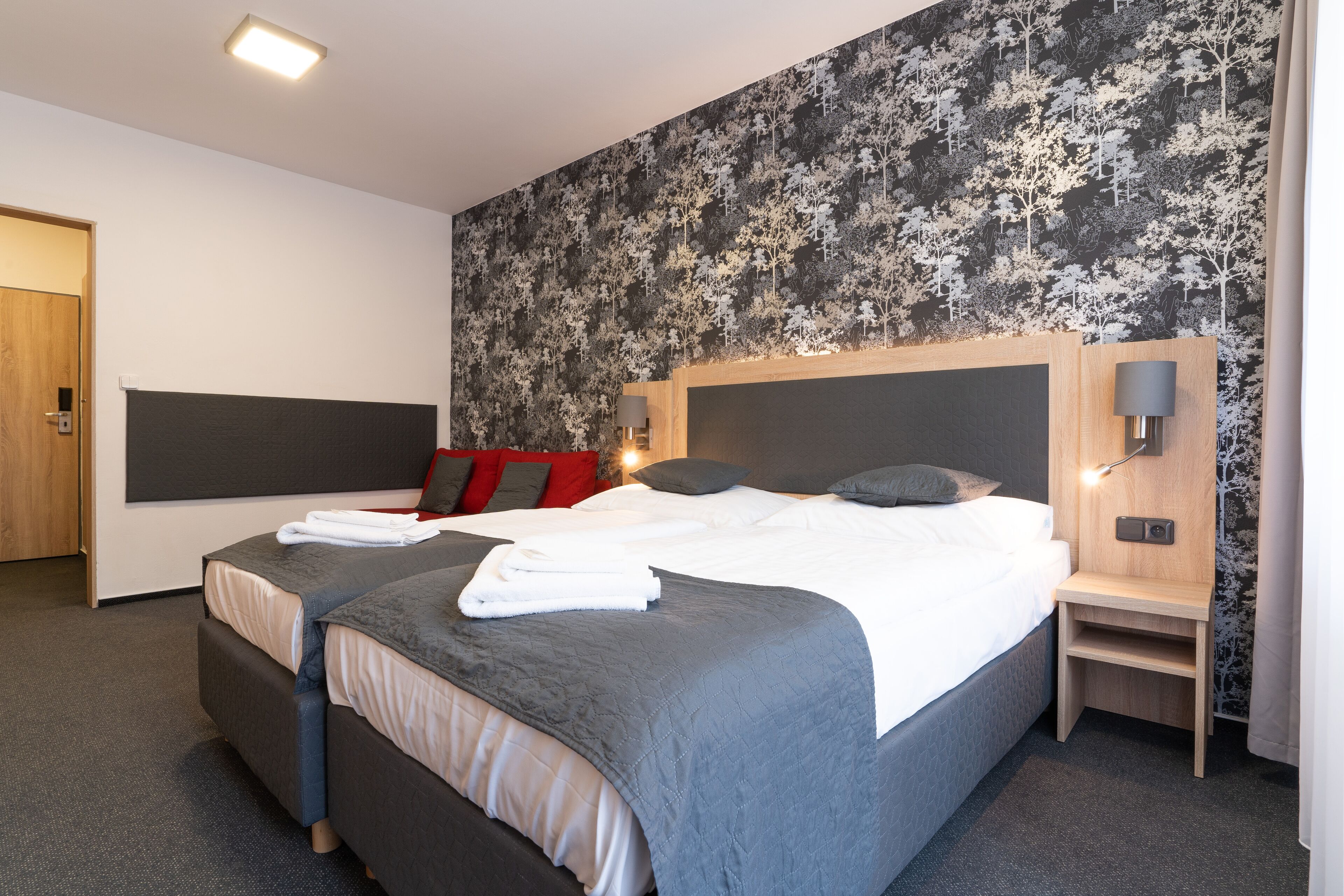A modern hotel room with a large bed, a bedside table, a nightstand, and a wall-mounted lamp.