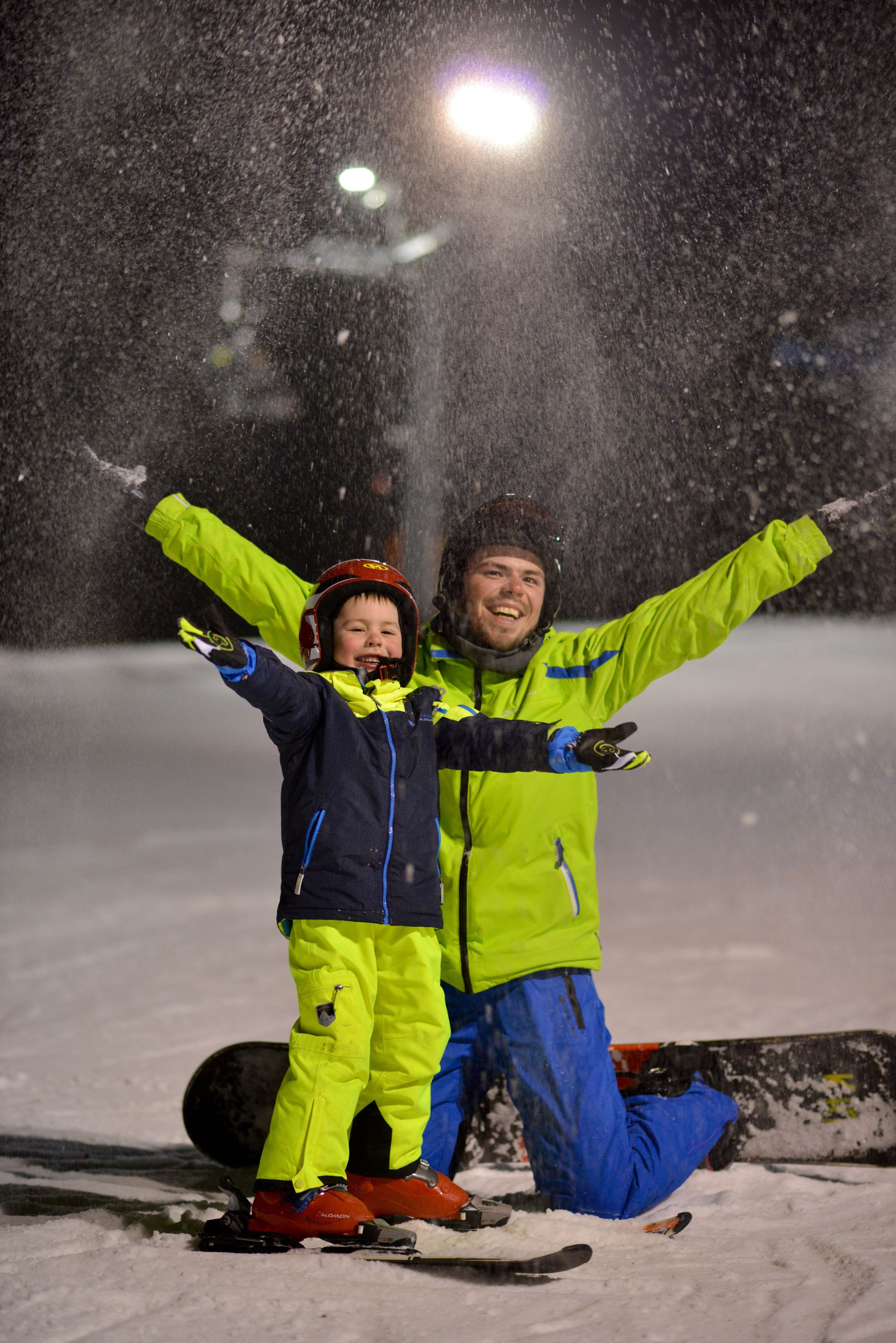 Deux personnes, vêtues de vêtements d’hiver, les bras levés, se tiennent sur la neige, avec une planche de snowboard à proximité.
