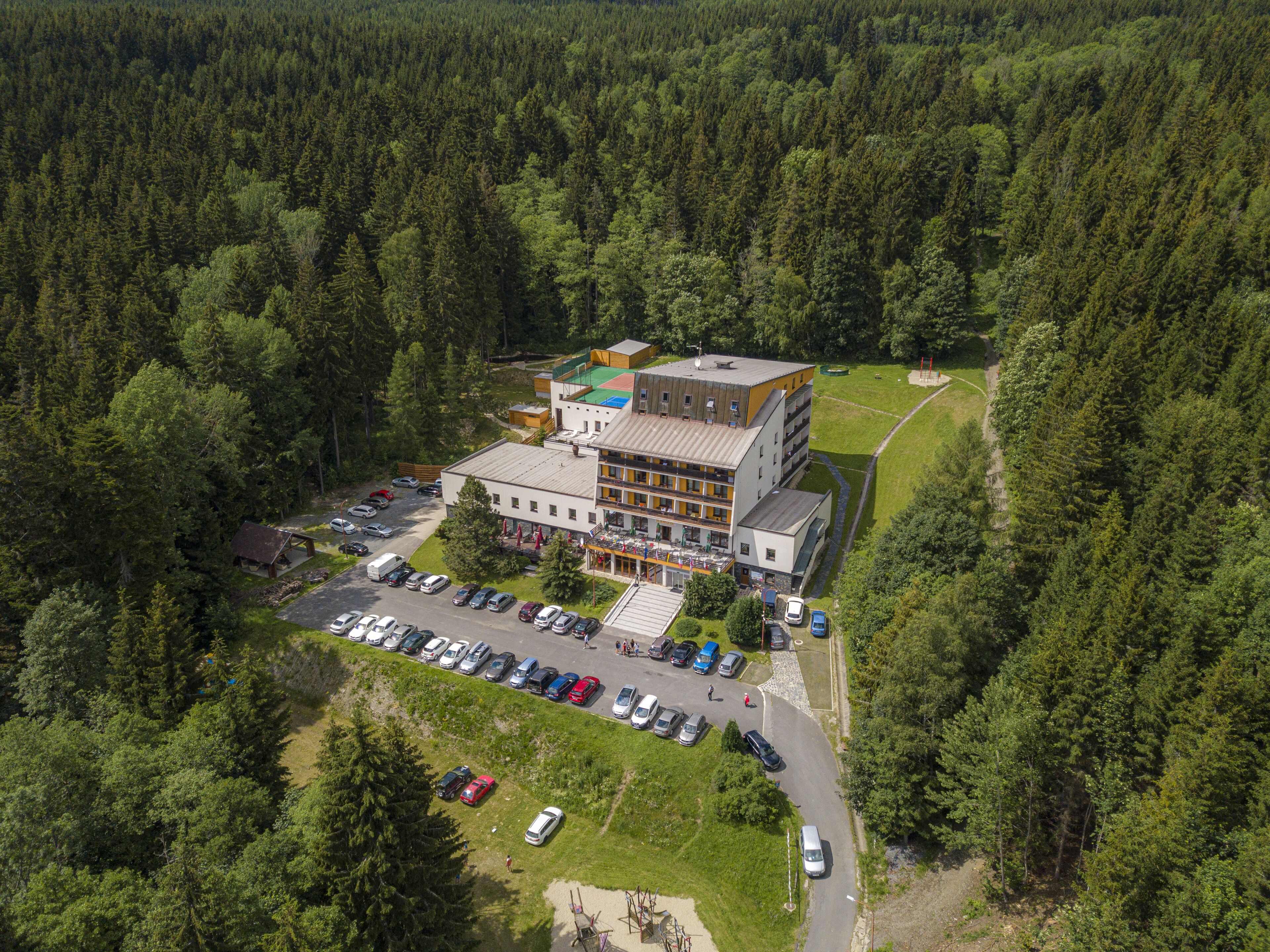 Vista aérea de um hotel de vários andares rodeado por floresta densa, com um estacionamento e uma pequena área externa.