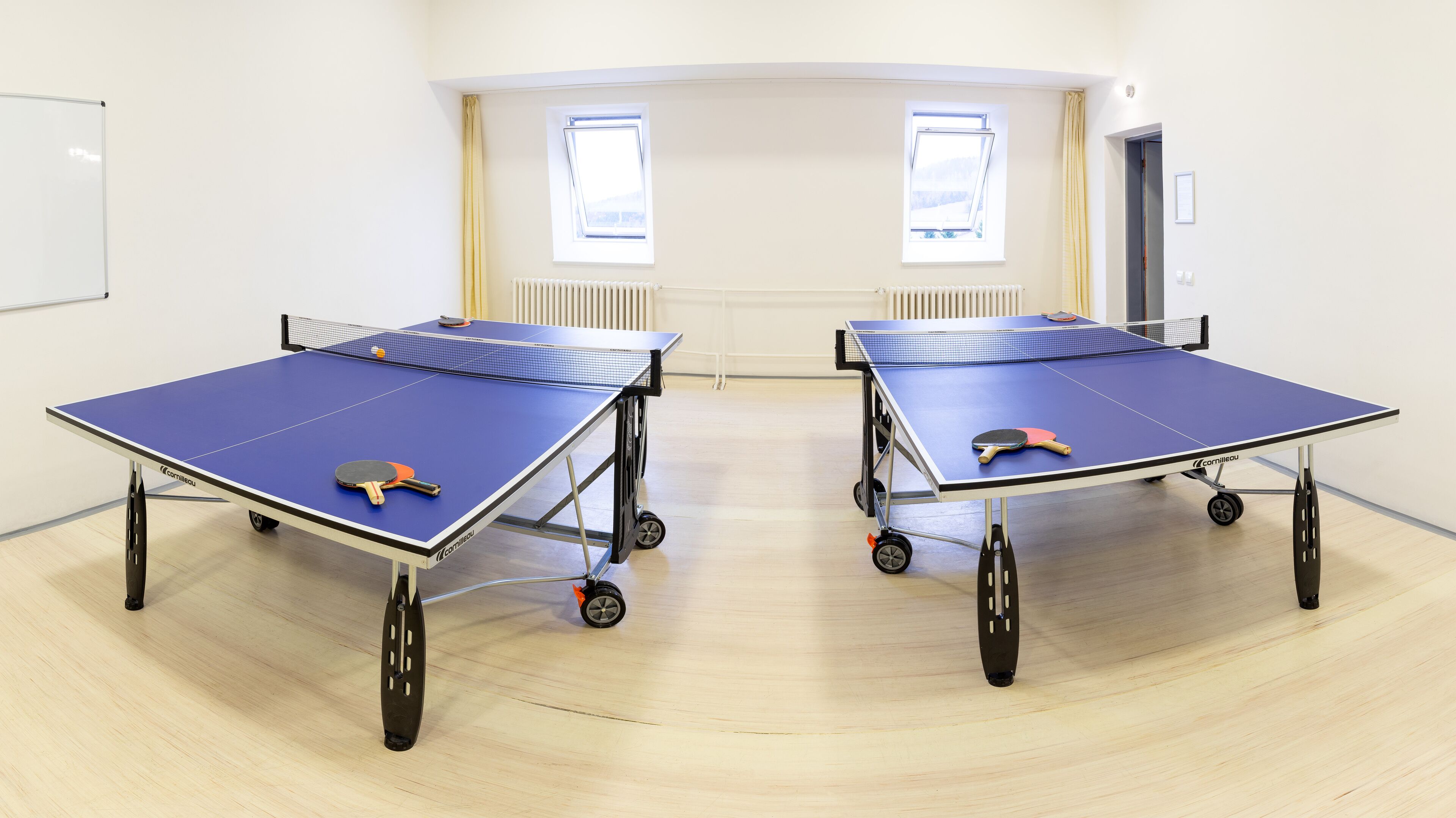 Dos mesas de ping pong en una habitación con suelo de madera clara y paredes blancas.