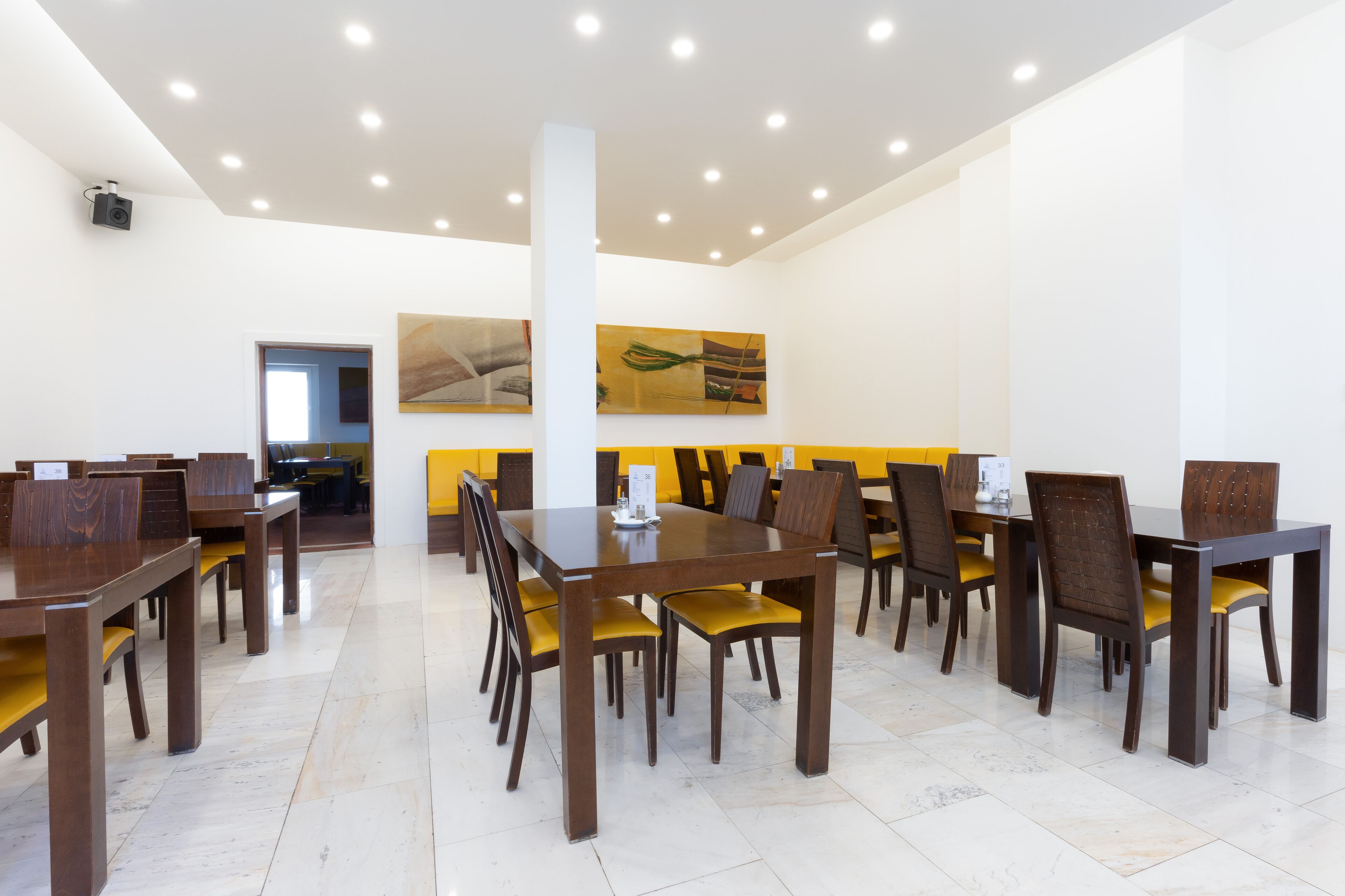 Ein geräumiger Essbereich mit mehreren Holztischen und -stühlen, einer weißen Decke mit Einbaustrahlern und einer gelben Akzentwand mit Kunstwerken.