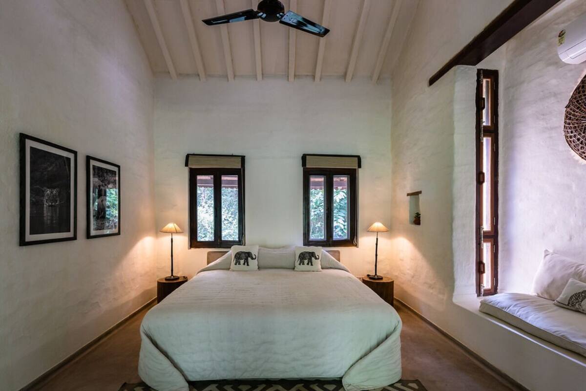 Un dormitorio con una cama grande, dos ventanas con contraventanas, un ventilador de techo y cuadros en la pared.