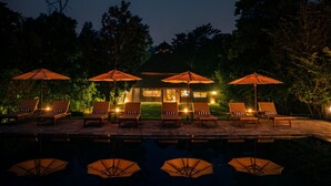 A nighttime outdoor seating area with lounge chairs and umbrellas, a pool, and a building with illuminated windows. - Tiger Tops Karnali Lodge (Thakudwara)