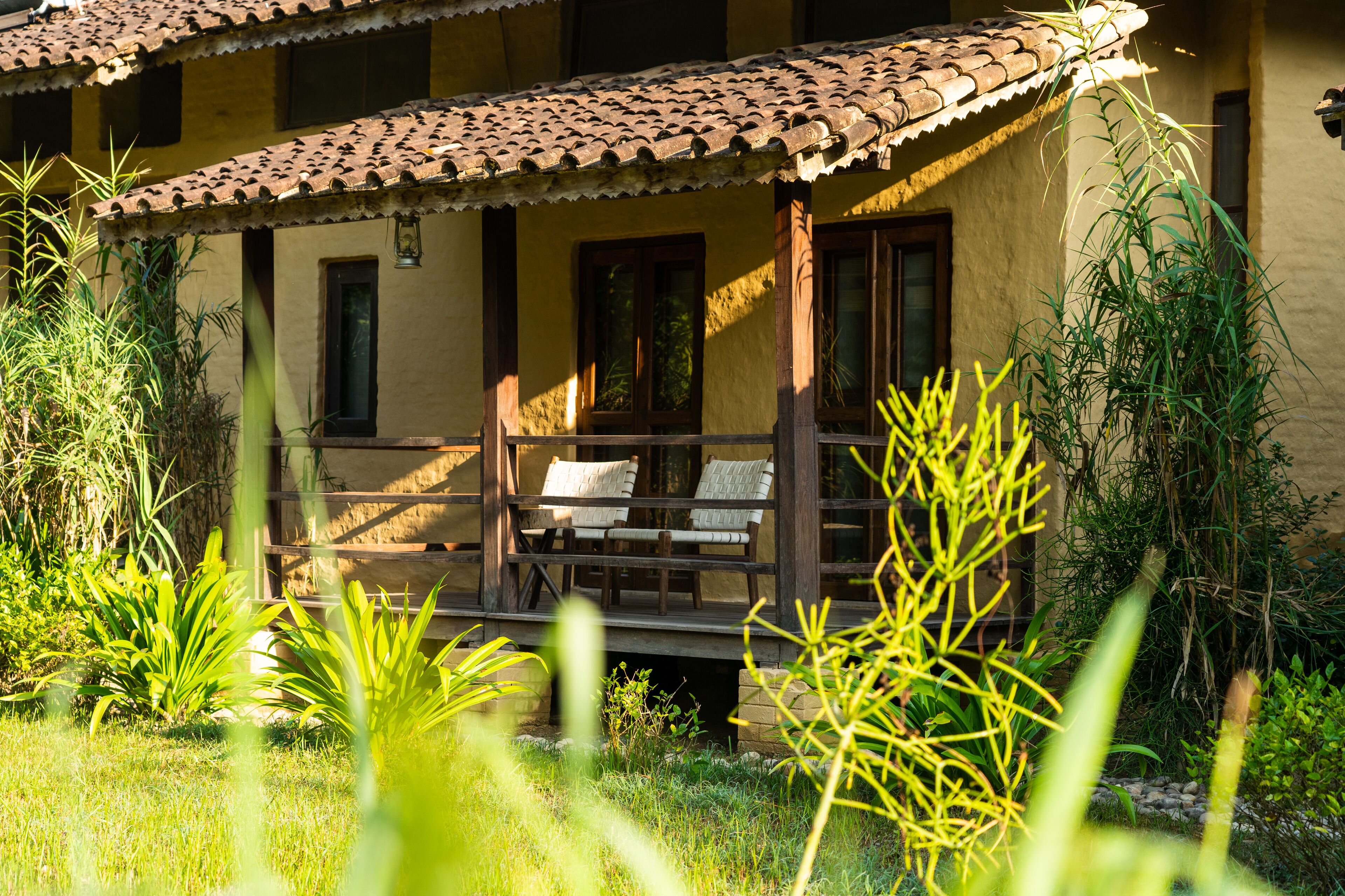 A building with a tiled roof, a wooden porch with two chairs, and surrounding greenery.