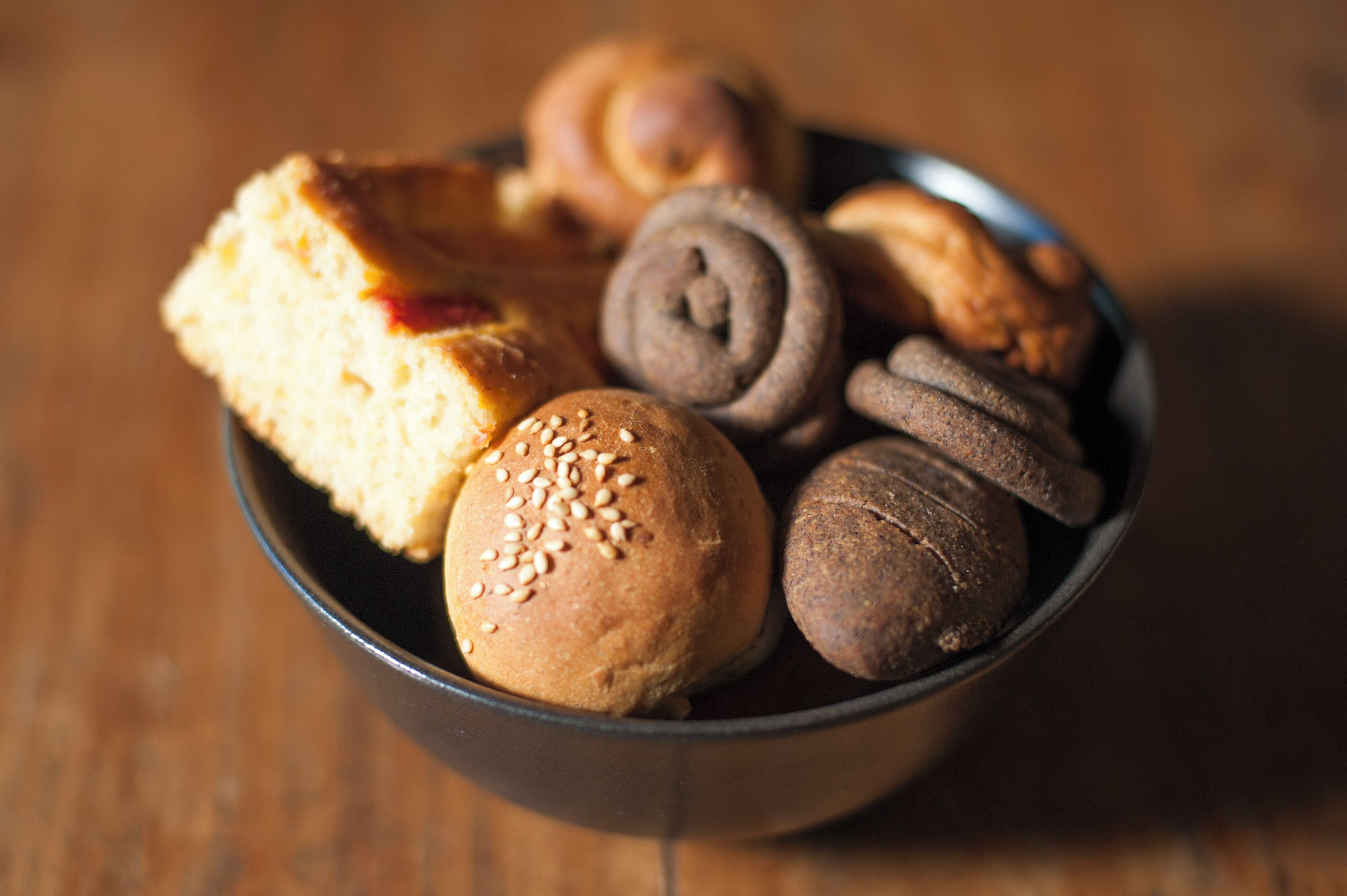 A bowl containing various baked goods, including a slice of pie, a round bun with sesame seeds, and several chocolate rolls.