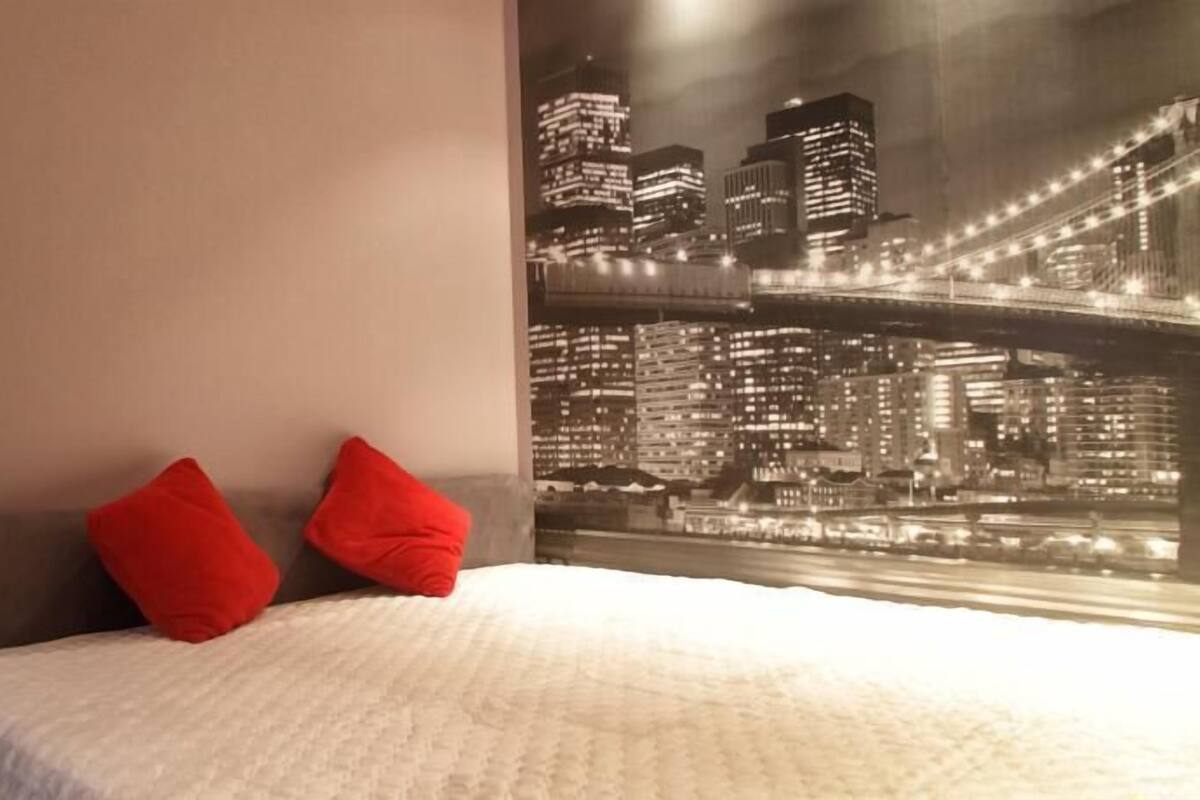 Een slaapkamer met een bed, twee rode kussens en een muurschildering van een stadsgezicht.