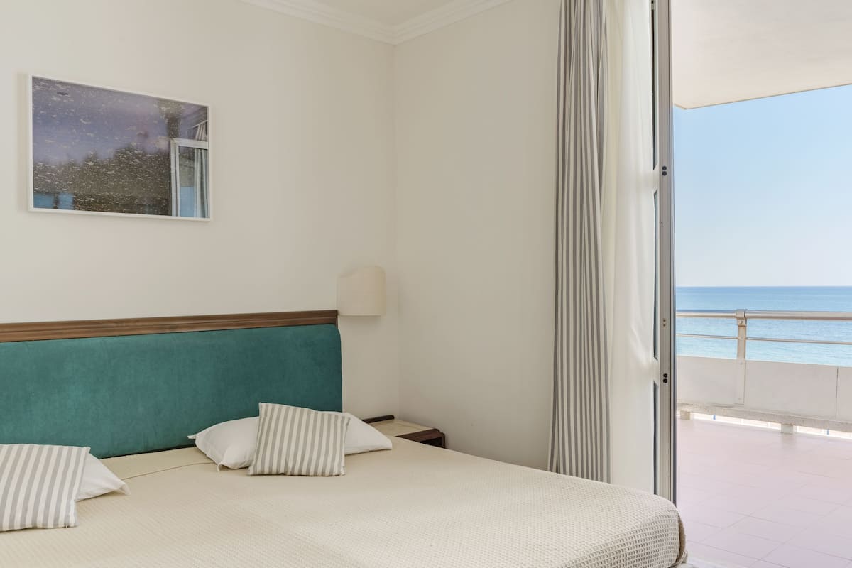 A hotel room with a bed, pillows, a nightstand, a wall-mounted lamp, a painting, and a view of the sea through a balcony door.